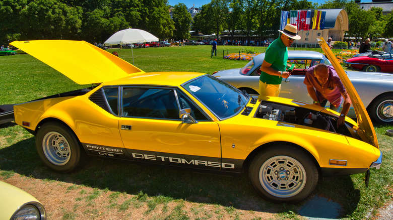 Two men looking under the hood of a yellow De Tomaso Pantera at a car show.