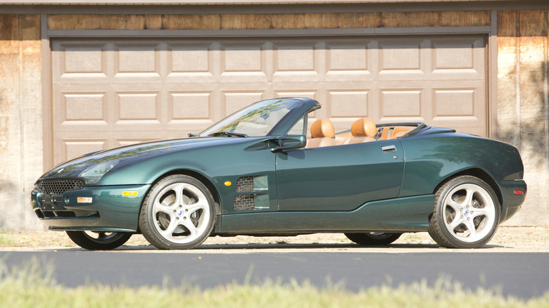A green Qvale Mangusta convertible with the top down parked in front of a garage.