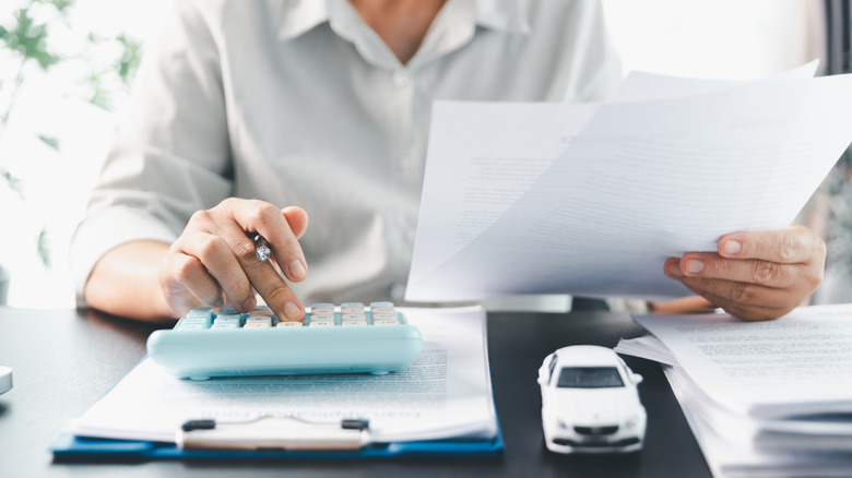 A person holds a few papers and crunches numbers on a calculator with a toy car nearby.