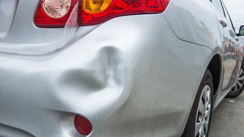 A silver car parked in a lot with a large dent on its right-rear fender