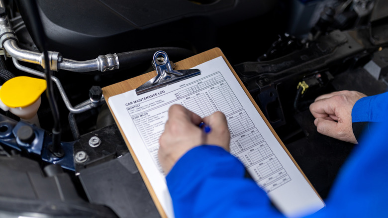 A mechanic completes detailed records on a clipboard in front of a car's engine bay