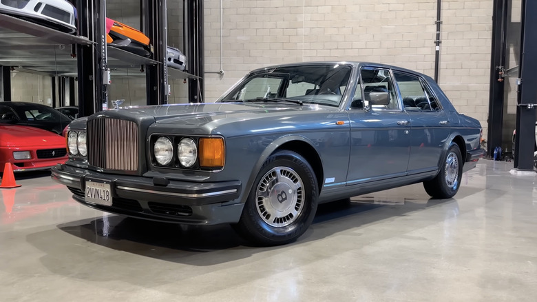 A depreciated blue Bentley sits parked in a garage with other cars on multi-tiered storage in the background