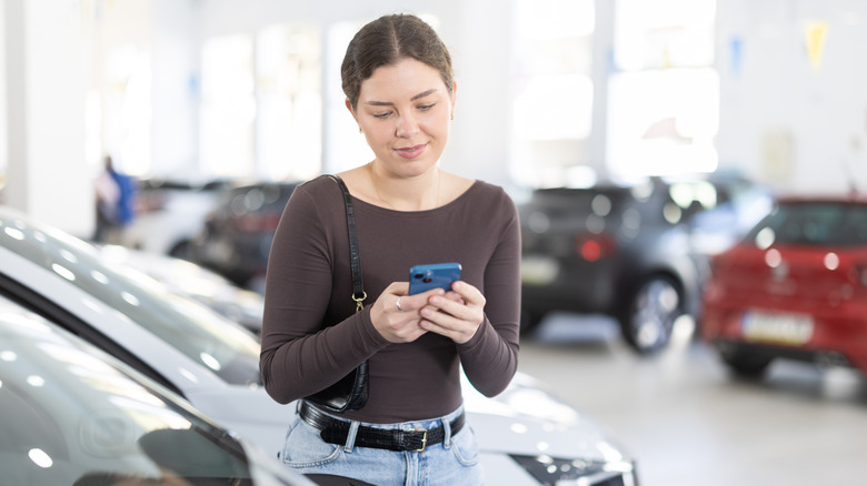 A person studies their phone at a car dealership with various vehicles in the background