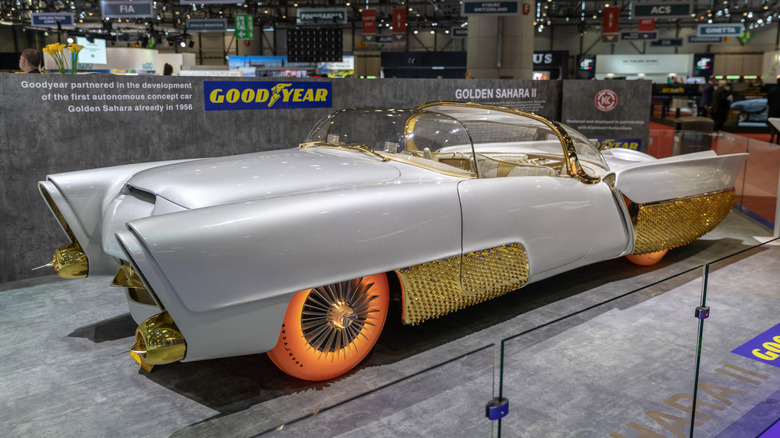 Front-three-quarter view of the Golden Sahara II custom concept car on an exhibit stage, featuring a pearl white body, gold-textured side skirts, a twin-bubble canopy, and illuminated orange translucent Goodyear tires.