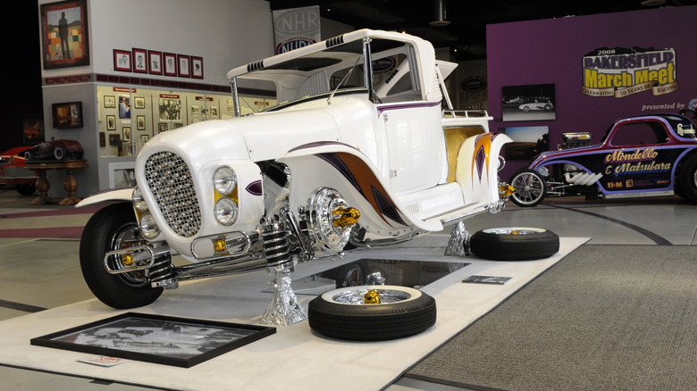 Front-three-quarter view of the white 1957 Ala Kart custom pickup truck with purple and gold accent scallops, displayed on stands inside a museum with its wheels unmounted on the floor.