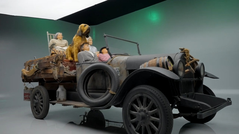 Front-three-quarter view of the rustic Beverly Hillbillies truck prop on a green screen studio set, loaded with family mannequins, tied-down household goods, and a large hound dog statue.
