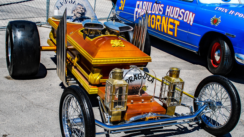 Outdoor front-three-quarter view of the Drag-U-La coffin dragster, featuring a gold-finished coffin body, exposed engine, large rear slick tires, front brass lanterns, and a vampire driver mannequin under a bubble canopy.