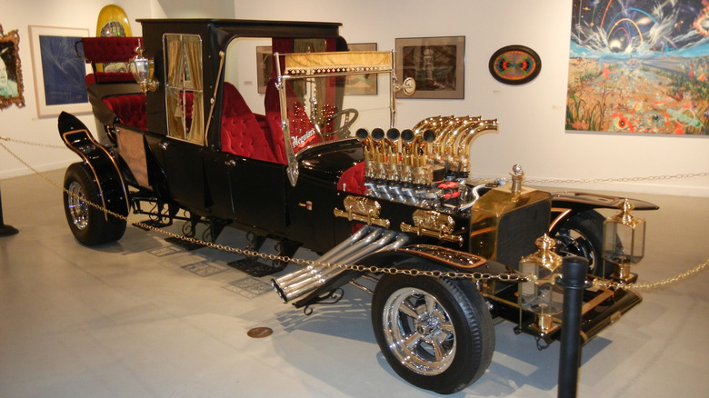 Front-three-quarter view of the Munster Koach custom hot rod in a gallery setting, featuring a long black chassis, brass organ-pipe engine headers, red velvet button-tufted seats, and brass lantern headlights.