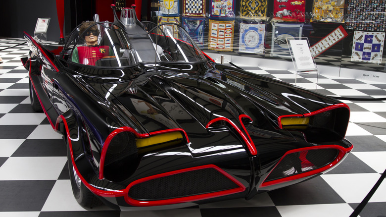 Front-three-quarter view of the 1966 Batmobile on display on a black-and-white checkered floor, featuring high-gloss black paint outlined in bright red pinstriping, twin Plexiglas bubble domes, and a Robin mannequin seated in the cockpit.