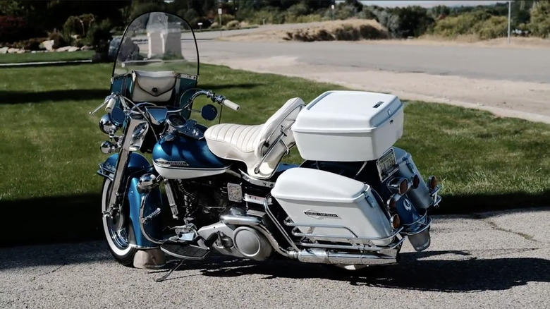 A blue and white 1965 Harley-Davidson FLH Electra Glide parked in an asphalt lot beside green grass.