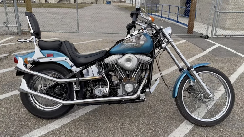 A blue 1984 Harley-Davidson FXST Softail with black and chrome accents parked on an asphalt lot with white stripes and a fence in the background.