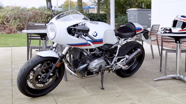 A 2020 BMW R nineT Racer parked outside on a paver laid patio next to a matching helmet and set of gloves that are resting on a table.