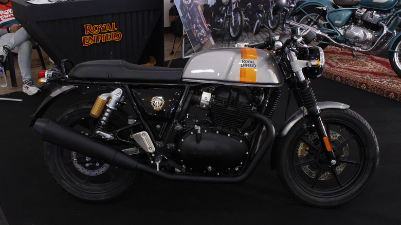 A 2025 Royal Enfield Continental GT 650 with black accents, a silver tank, and an orange stripe sitting on a black floor in a motorcycle showroom.