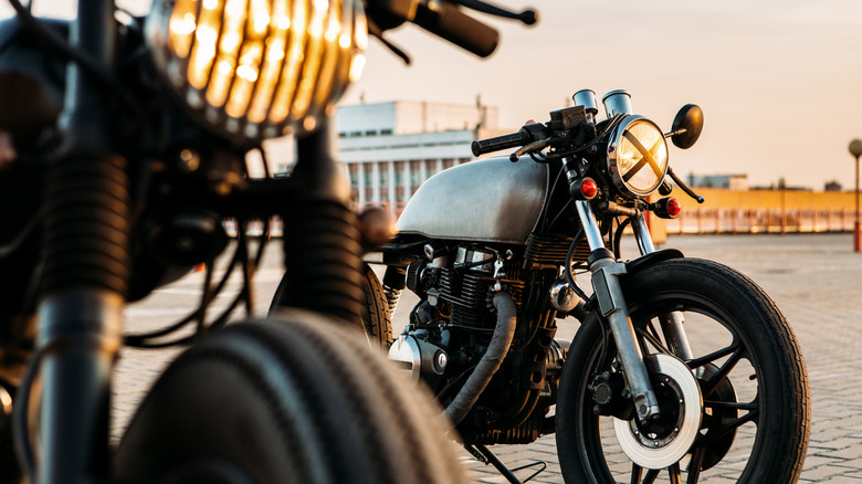 A pair of vintage cafe racers resting in the sunset with headlights on in a brick paved lot.