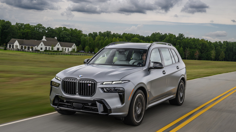 A gray BMW X7 drives past an open field with a rural home in the background