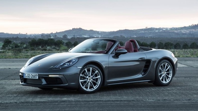 A Porsche 718 Boxster parked on pavement in front of green hills.