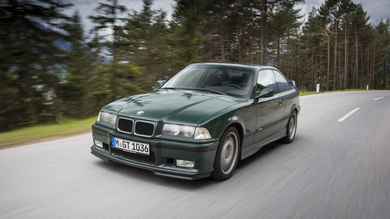 A green E36 BMW drives by some trees.