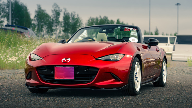 A red Mazda Miata sits parked on pavement with tall grass in the background