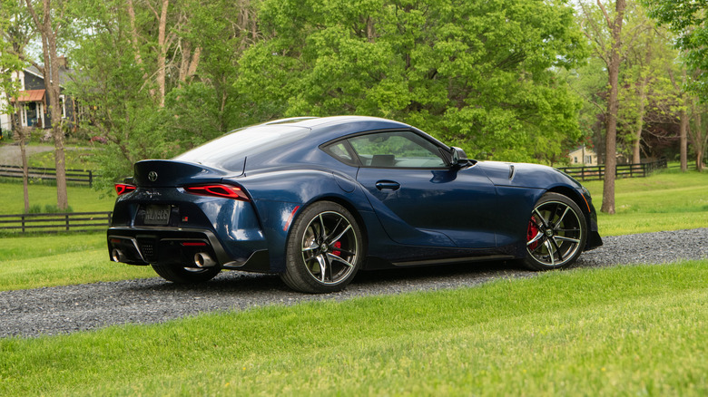 A blue Toyota Supra parked on a gravel path with trees and a fence in the background