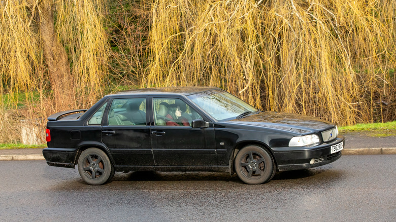 A dark-colored Volvo S70 driving on the road near woods.