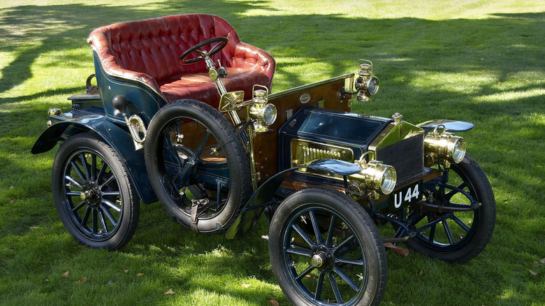 An open-topped Rolls-Royce sits in the shade on a lawn with its red leather seat sitting up high.