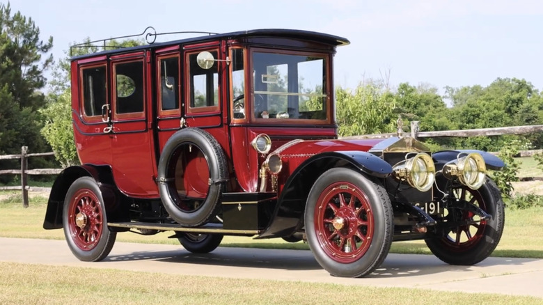 A red Rolls-Royce limousine sits on a small road near a field with a full-sized spare on the door.