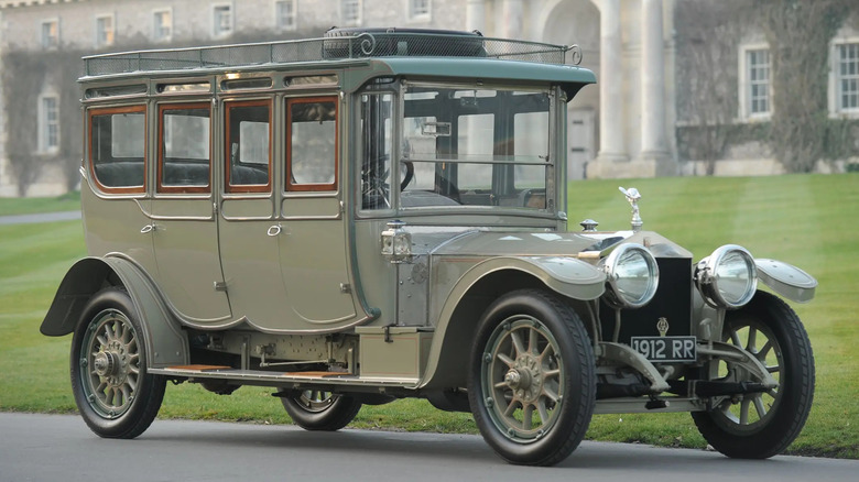 A striking Rolls-Royce Silver Ghost sits near a manicured lawn in front of an impressive stone building.