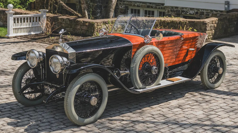 A vintage Rolls-Royce sits in a brick driveway outside a home, with wooden bodywork gleaming.