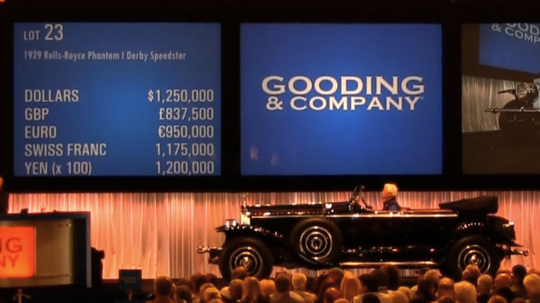 A black Rolls-Royce Phantom I Derby Speedster sits on a stage in an auction with screens behind and a crowd gathered to watch.