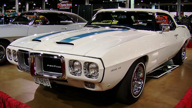 A white 1969 Pontiac Firebird Trans Am convertible with blue racing stripes on display at an indoor car show