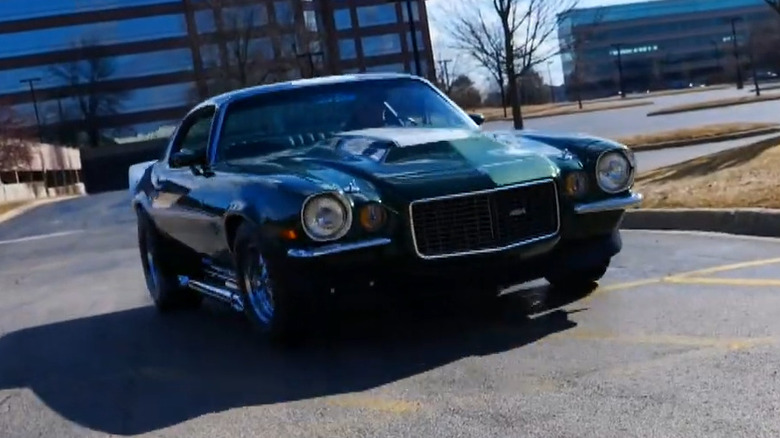 A dark green 1970 Chevrolet Camaro Baldwin-Motion Phase III with custom cowl hood and side exhaust, driving outdoors in a parking lot