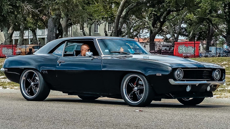 A black 1969 Chevrolet Camaro ZL1 coupe cruising on an open road with custom wheels and a cowl induction hood