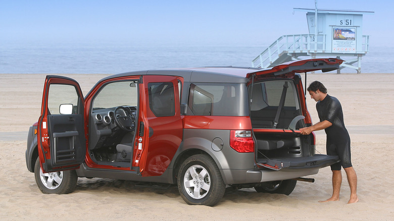 A sunset-lit beach scene featuring an orange and gray Honda Element with its rear clamshell tailgate and side suicide doors fully open.