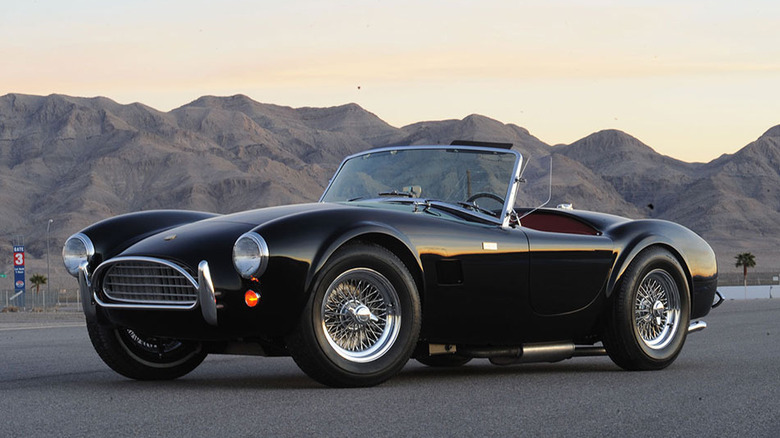 A black Shelby Cobra 289 "Slab-side" roadster with chrome wire wheels, parked on a racetrack at dusk.