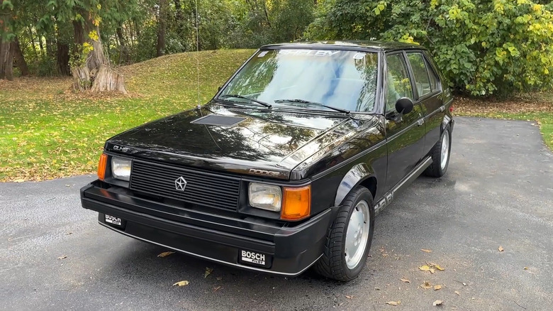 A black 1986 Dodge Shelby Omni GLHS parked in a driveway. The image captures the signature "GLHS" decal on the hood, the front air dam with Bosch Pilot fog lights, and the silver "Centurion" wheels.