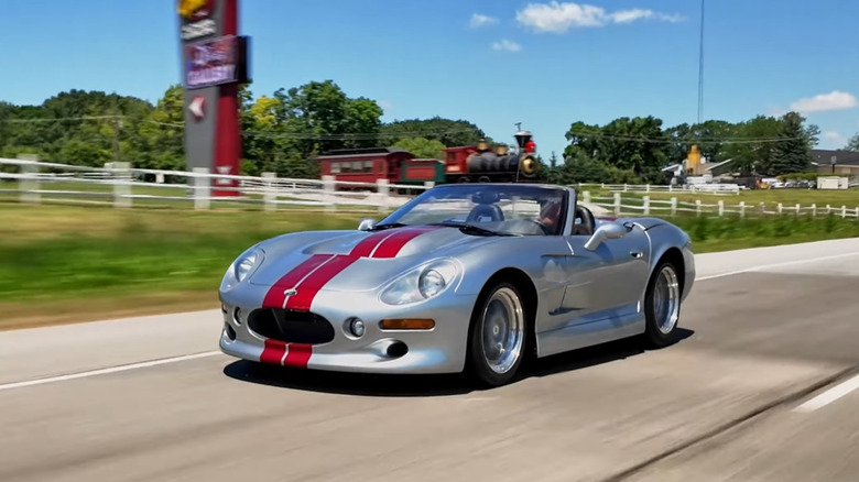 A silver 1999 Shelby Series 1 with dual red racing stripes in motion on a highway.