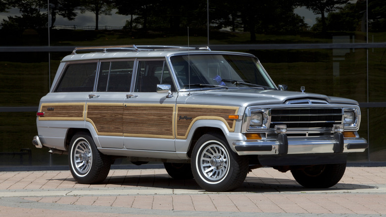 A silver 1989 Jeep Grand Wagoneer parked in front of a dark, modern glass building, prominently displaying its iconic wood-grain side paneling.