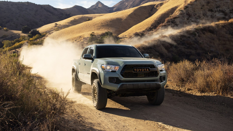 A light-colored 2023 Toyota Tacoma pickup truck kicking up dust while driving along a dirt trail through golden, sunlit hills.
