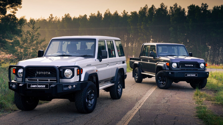 A pair of 2026 Toyota Land Cruiser 70 Series vehicles—a four-door SUV and a two-door pickup truck—parked on a quiet, tree-lined road during a golden sunset.