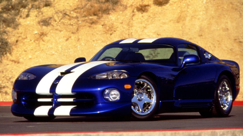 A low, front-quarter angle of a classic blue 1997 Dodge Viper GTS with white racing stripes, parked on an outdoor tarmac against a bright, hazy sky.