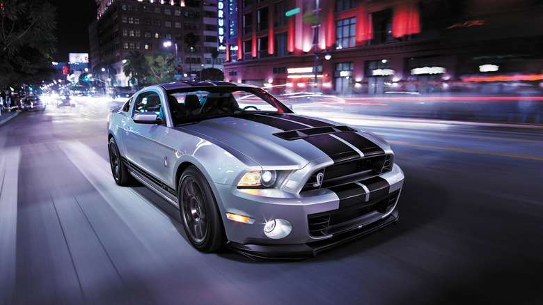 A silver Ford Mustang Shelby GT500 with black racing stripes captured in a long-exposure shot driving through a vibrant, neon-lit city street at night.