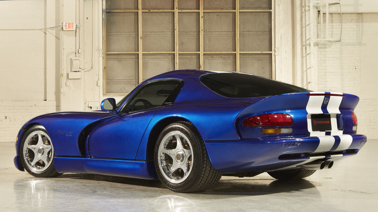 A rear 3/4 view of a vibrant blue Dodge Viper RT/10 with white racing stripes, parked in a clean, industrial-style warehouse with large windows.