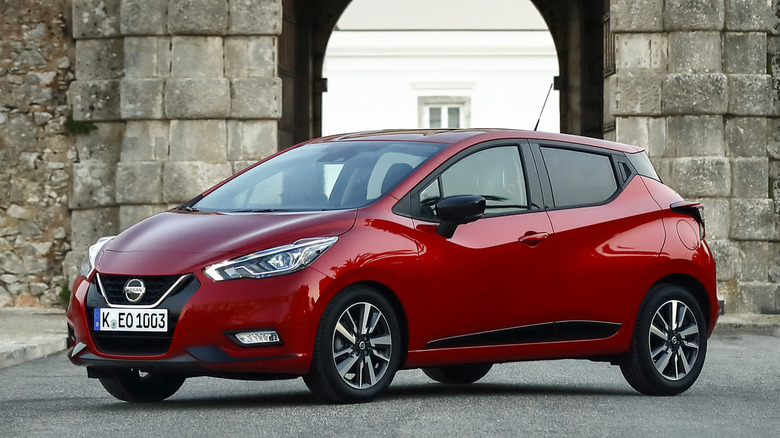 Bright red Nissan Micra hatchback is parked on a paved road in front of stone-walled entrance.