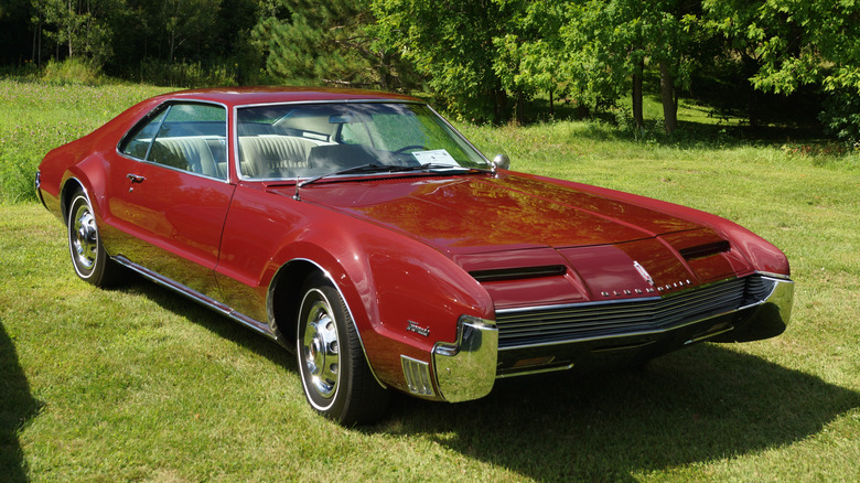 Red first-generation Oldsmobile Toronado coupe right front three-quarter view parked on grass with trees in background