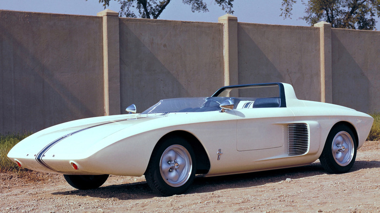The white 1962 Ford Mustang I mid-engine concept speedster parked outdoors on gravel, highlighting its wedge front end, blue racing stripe, integrated roll bar, and deep side air intake vents.