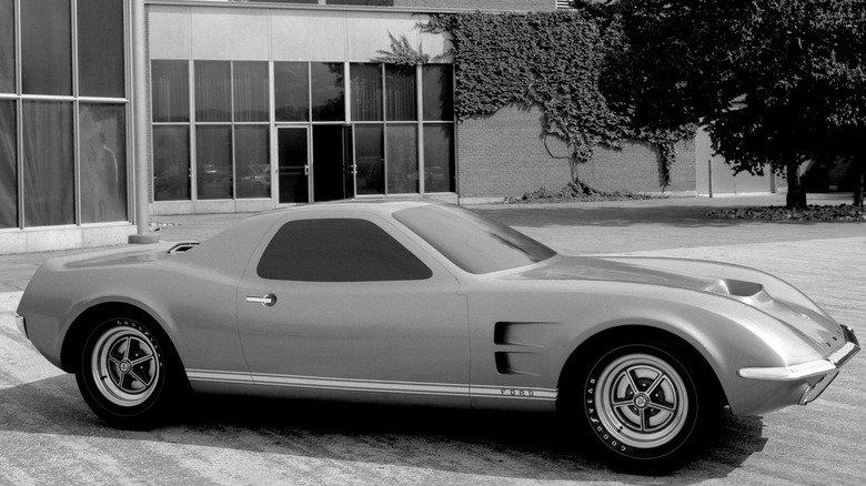 A black-and-white photo of the 1967 Ford Mach 2 mid-engine concept coupe parked outside the Ford styling building, showing the car's long rear deck, front fender vents, and classic mag-style wheels.