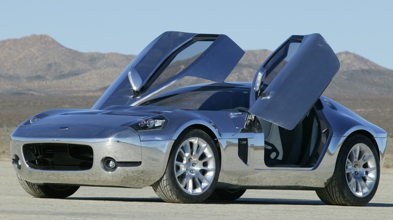 The polished bare-aluminum 2004 Ford Shelby GR-1 concept car parked on a desert dry lake bed with its butterfly doors raised upward.
