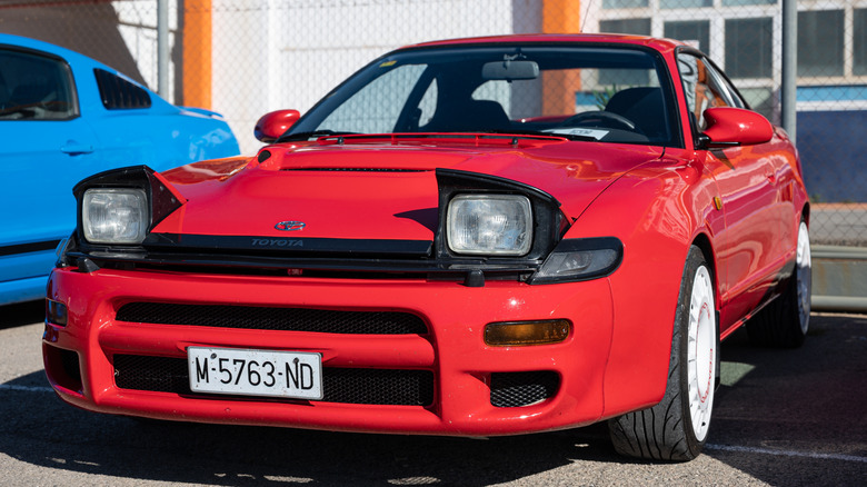 The front three-quarter view of a red fifth-generation Toyota Celica All-Trac Turbo (ST185) parked in front of a fenced building next to an S550 Mustang