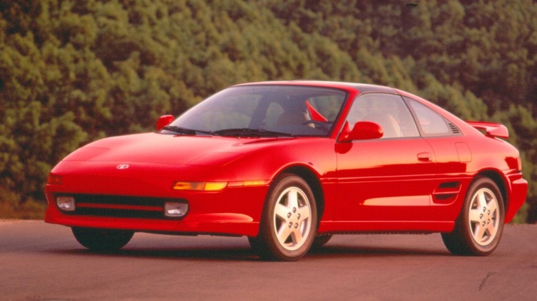The front three-quarter view of a bright red second-generation Toyota MR2 Turbo coupe parked on asphalt, showcasing its pop-up headlamp front clip, side engine air intake, five-spoke alloy wheels, and rear wing.
