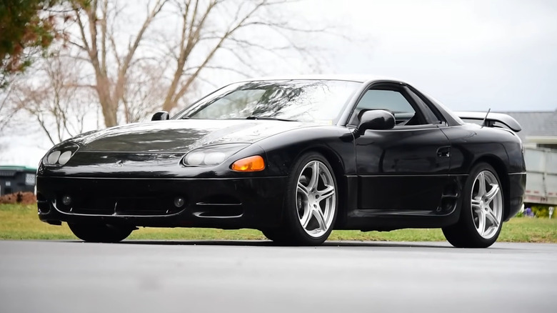 A Low-angle front three-quarter shot of a black second-generation Mitsubishi 3000GT parked on asphalt, showing its fixed projector headlights, side strakes, and silver aftermarket wheels.